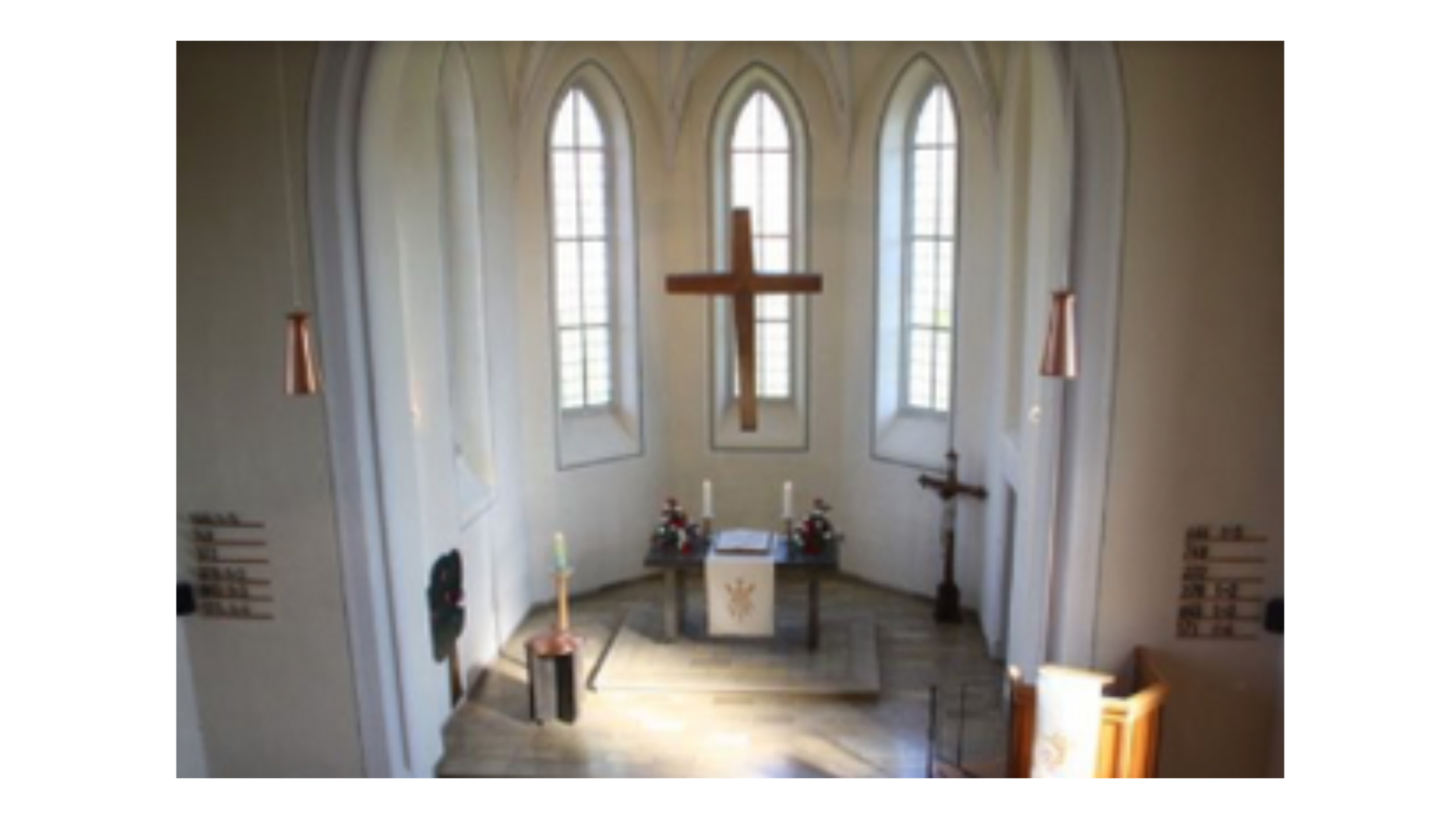 Kircheninnenraum mit einem großen Holzkreuz vor drei hohen Fenstern.