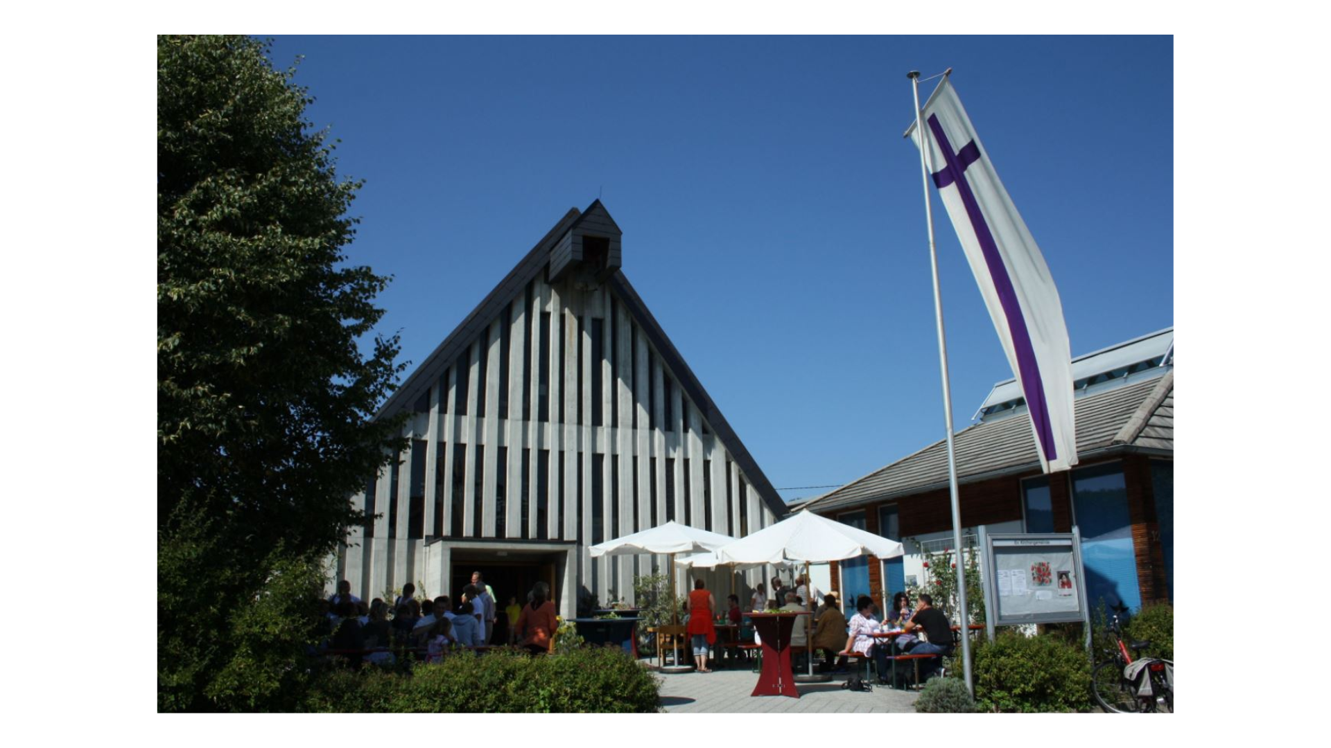 Moderne Kirche mit vertikalen Lamellen, davor Festzelt und Fahne mit Kreuz.