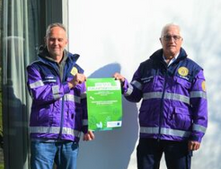 Zwei Männer in lila Jacken halten ein grünes Plakat.